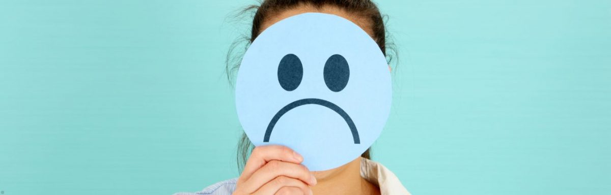 A woman holding a paper sad face in front of her head. She's standing against a teal background.