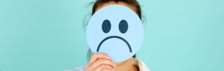 A woman holding a paper sad face in front of her head. She's standing against a teal background.