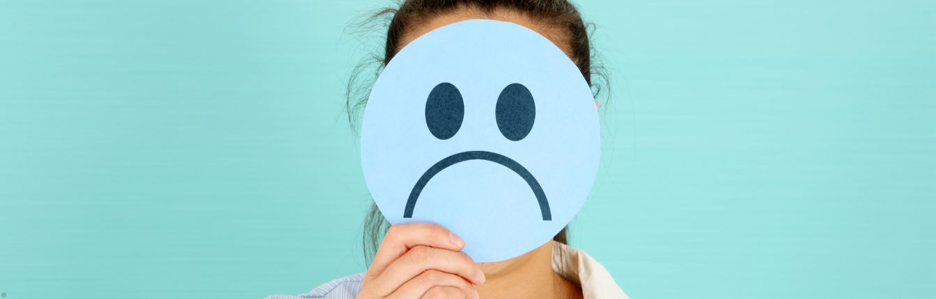 A woman holding a paper sad face in front of her head. She's standing against a teal background.