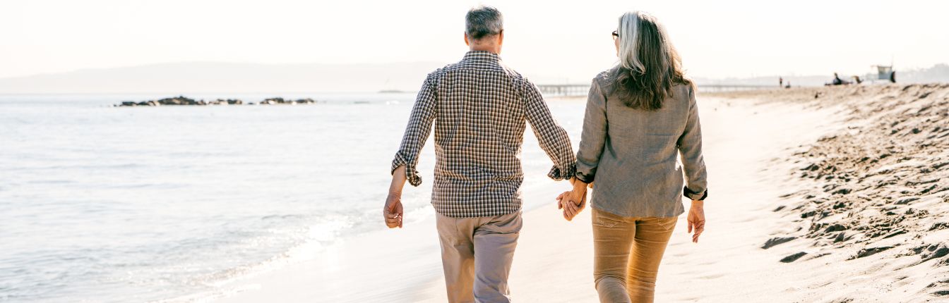 An older couple, holding hands and walking on a beach.