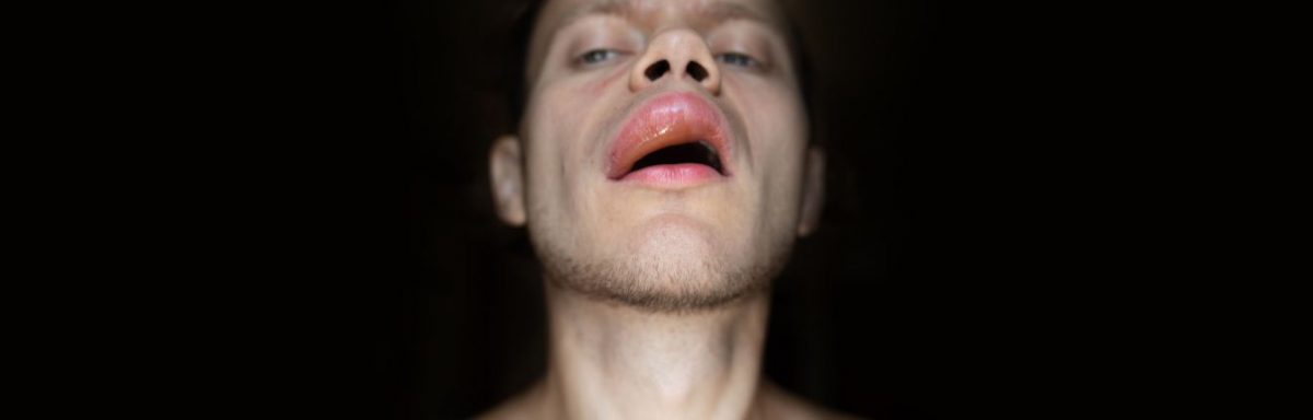 A man's face with very swollen lips, against a black background.