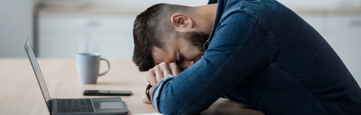 A man laying his head down on a table, in front of a laptop.