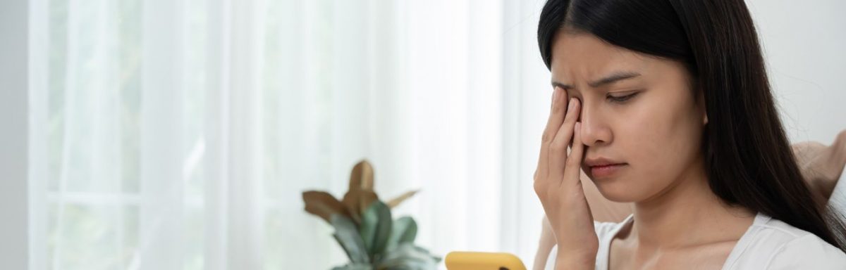 A young woman rubbing her eye while looking at her cell phone.